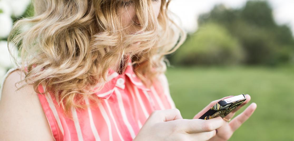 Somrigt klädd kvinna med lockigt hår framför ansiktet tittar ner på mobilen hon håller i sina händer.