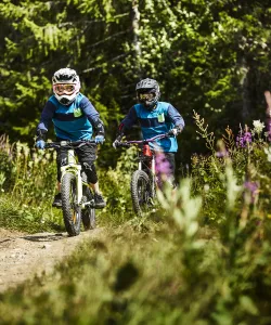 Två barn på cyklar i sommarnatur. De bär hjälm och skyddskläder.