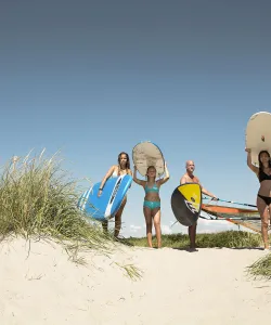 Tre kvinnor och en man med surfingbrädor på sandstrand.