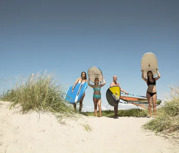 Tre kvinnor och en man med surfingbrädor på sandstrand.