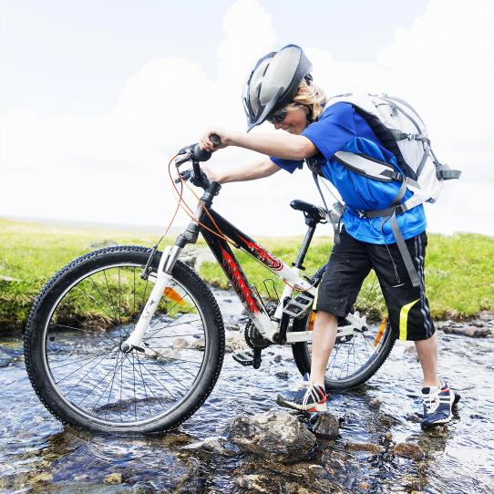 Kvinna leder sin cykel i en grunt vattendrag. Hon bär hjälm och ryggsäck.