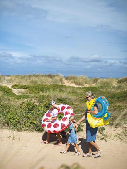 En vuxen och två barn bär badringar mota havet genom sanddyner.
