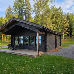 Mörk trästuga med veranda och utemöbler på gräsmatta, omgiven av höga träd.