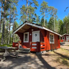 Röd trästuga med veranda och picknickbord i en solig tallskog.